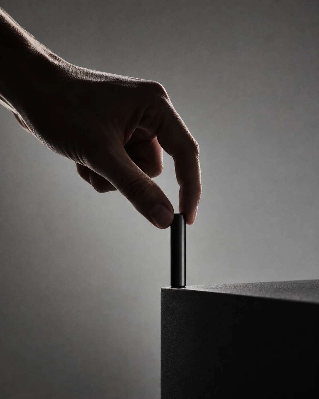 A hand balancing a small black cylinder at the edge of a graphite platform.